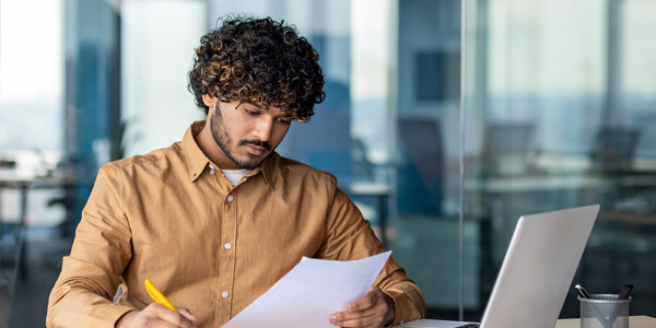 Young serious focused and thinking businessman paper work, successful hispanic man reviewing contract reports and invoices, sitting at desk inside office with laptop, checking and filling form