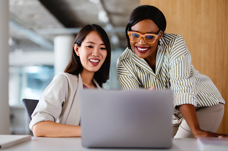 A woman with orange glasses shows another woman her laptop