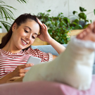 A woman looks her phone while elevating her injured leg that is wrapped in a cast