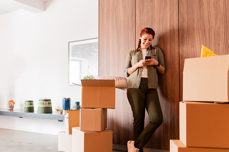 A woman smiles at her phone while stacks of moving boxes surround her