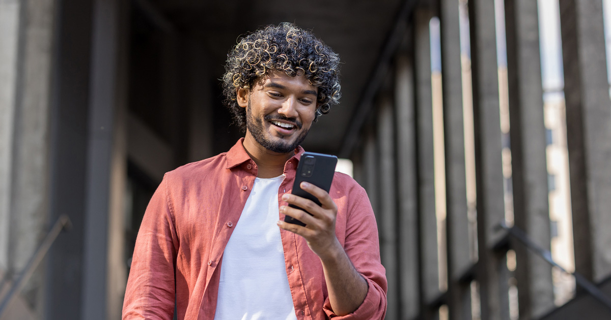 A man with a red button up shirt smiles at his phone under a bridge
