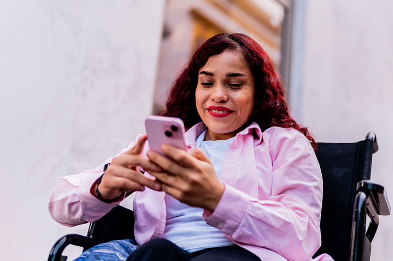 A woman wearing pink, types on her pink phone, while sitting in her wheelchair